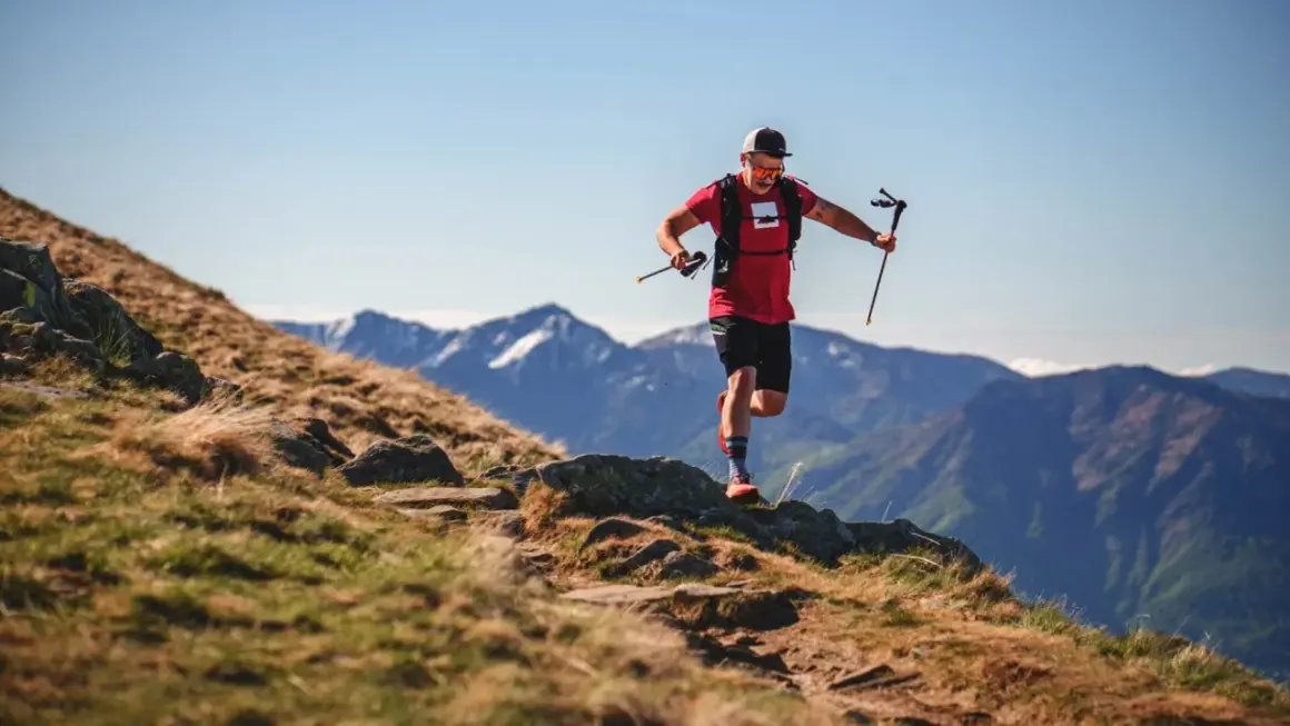Ein Mann im roten Shirt und schwarzer Hose läuft mit Stöcken einen Bergpfad entlang. Sein intensives anaerobes Training fordert ihn in der atemberaubenden Berglandschaft.