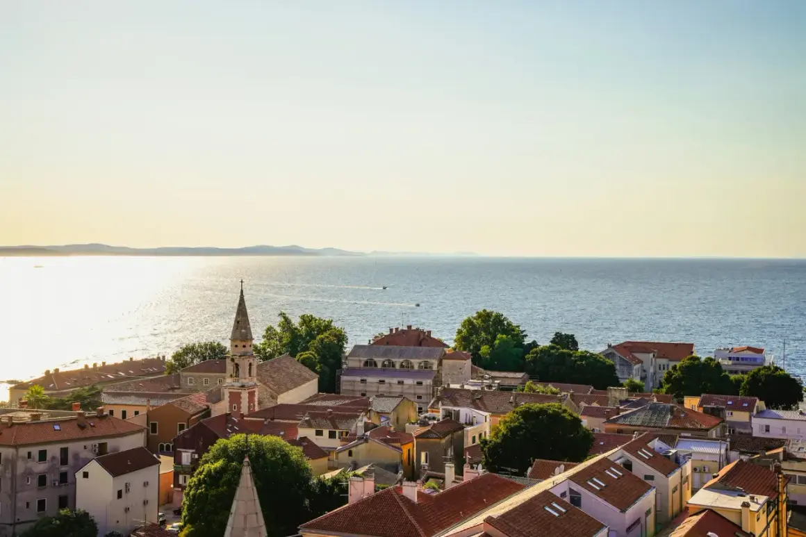 Widok na dachy Zadar i okolice, z kościelną wieżą górującą nad miastem i błękitnym morzem w tle.