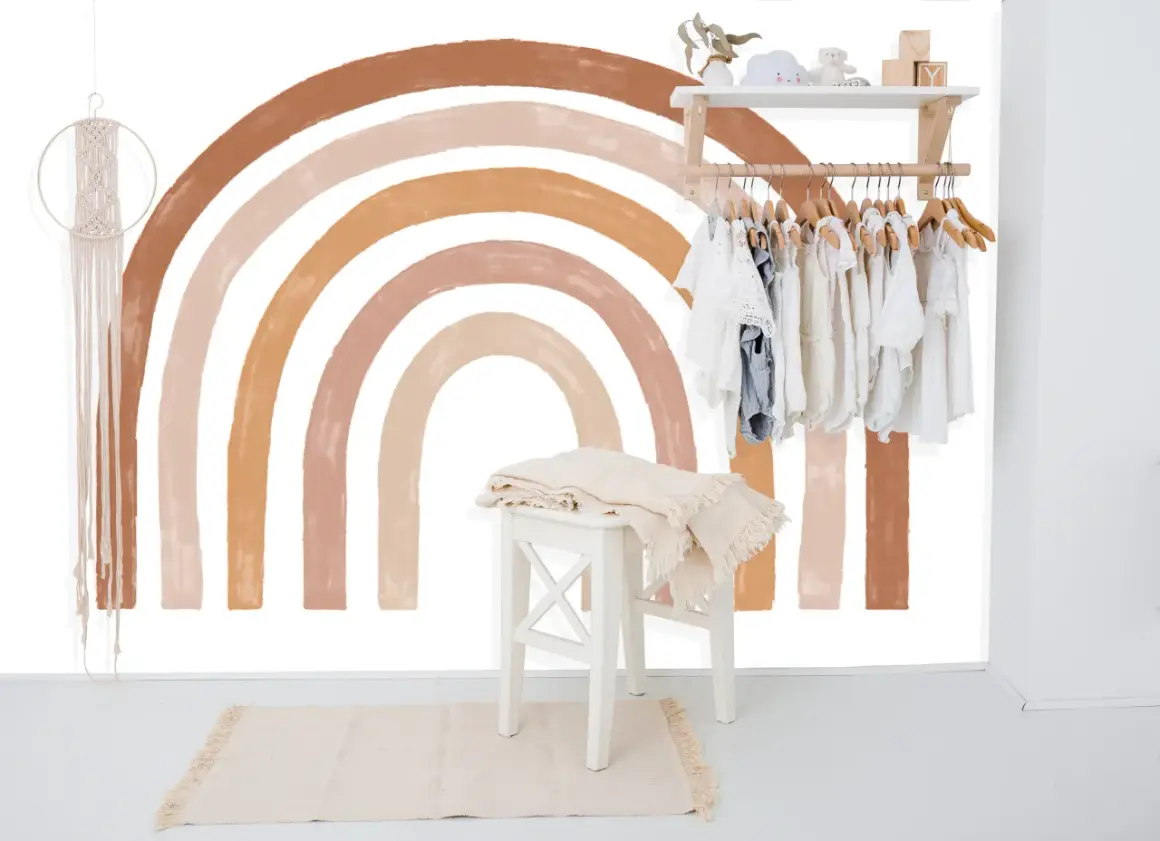 Un arc-en-ciel aux tons terreux est peint sur un mur blanc, orné d'une penderie d'enfants et d'une décoration murale en macramé.