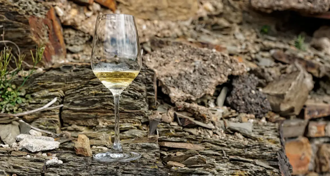 Ein Glas Weißwein steht auf schieferigem **Weinboden**. Die raue Gesteinslandschaft bildet einen starken Kontrast zum edlen Tropfen.