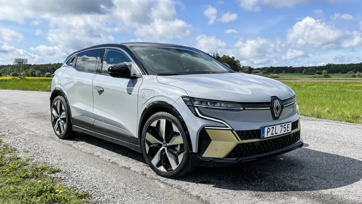 En vit Renault Megane E-Tech står parkerad på en grusväg med en gul och grön fält i bakgrunden. En del av en testkörning.