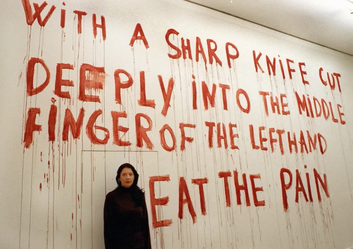 A woman stands before a wall with red, dripping text that reads "WITH A SHARP KNIFE CUT DEEPLY INTO THE MIDDLE FINGER OF THE LEFTHAND EAT THE PAIN." This is a reference to "spirit cooking.