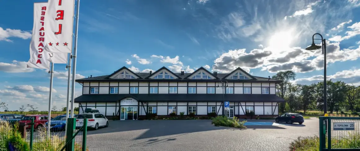 Hotel z białymi ścianami i czarnymi belkami, trzy gwiazdki na fladze, parking.