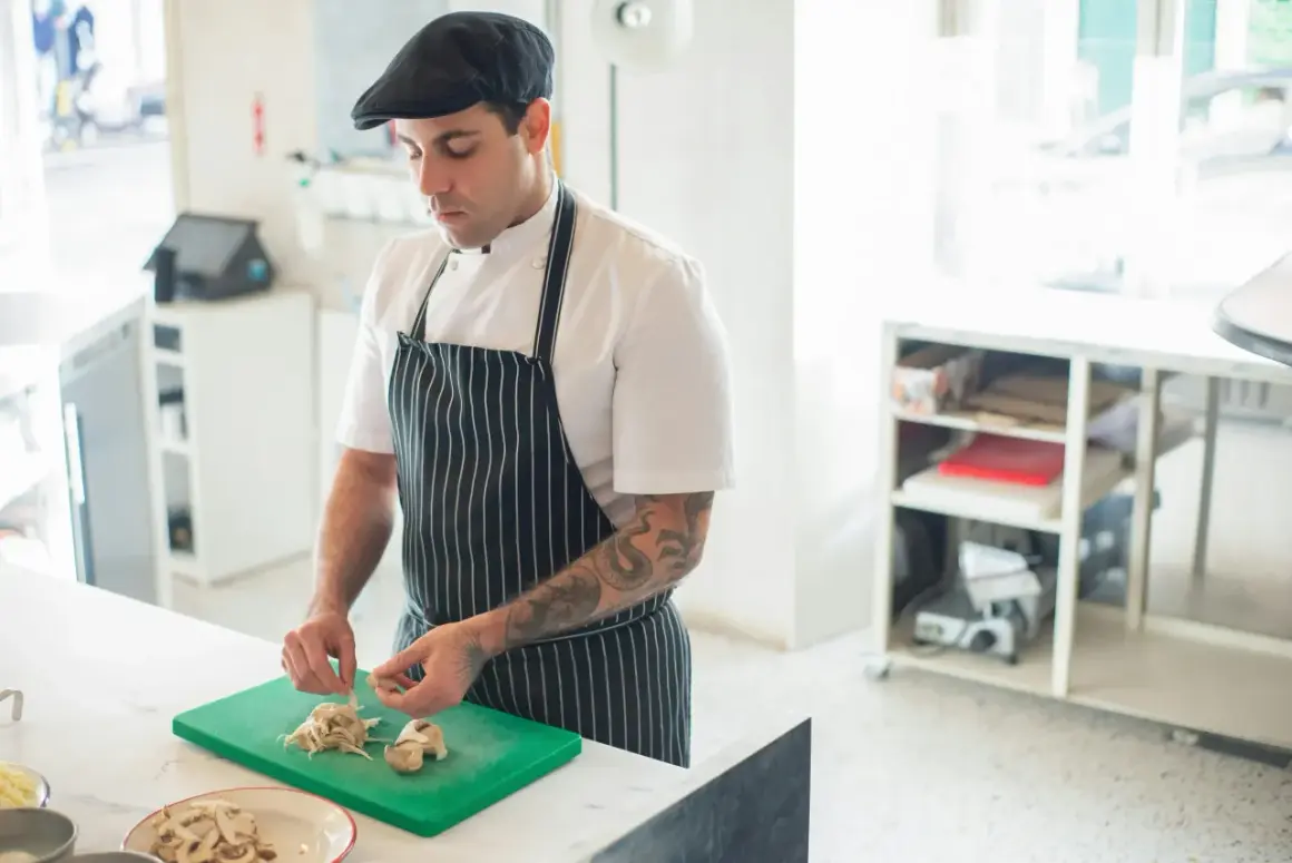 Chef con gorra y delantal a rayas, cortando champiñones en una tabla verde. Ideal para aprender a cocinar.