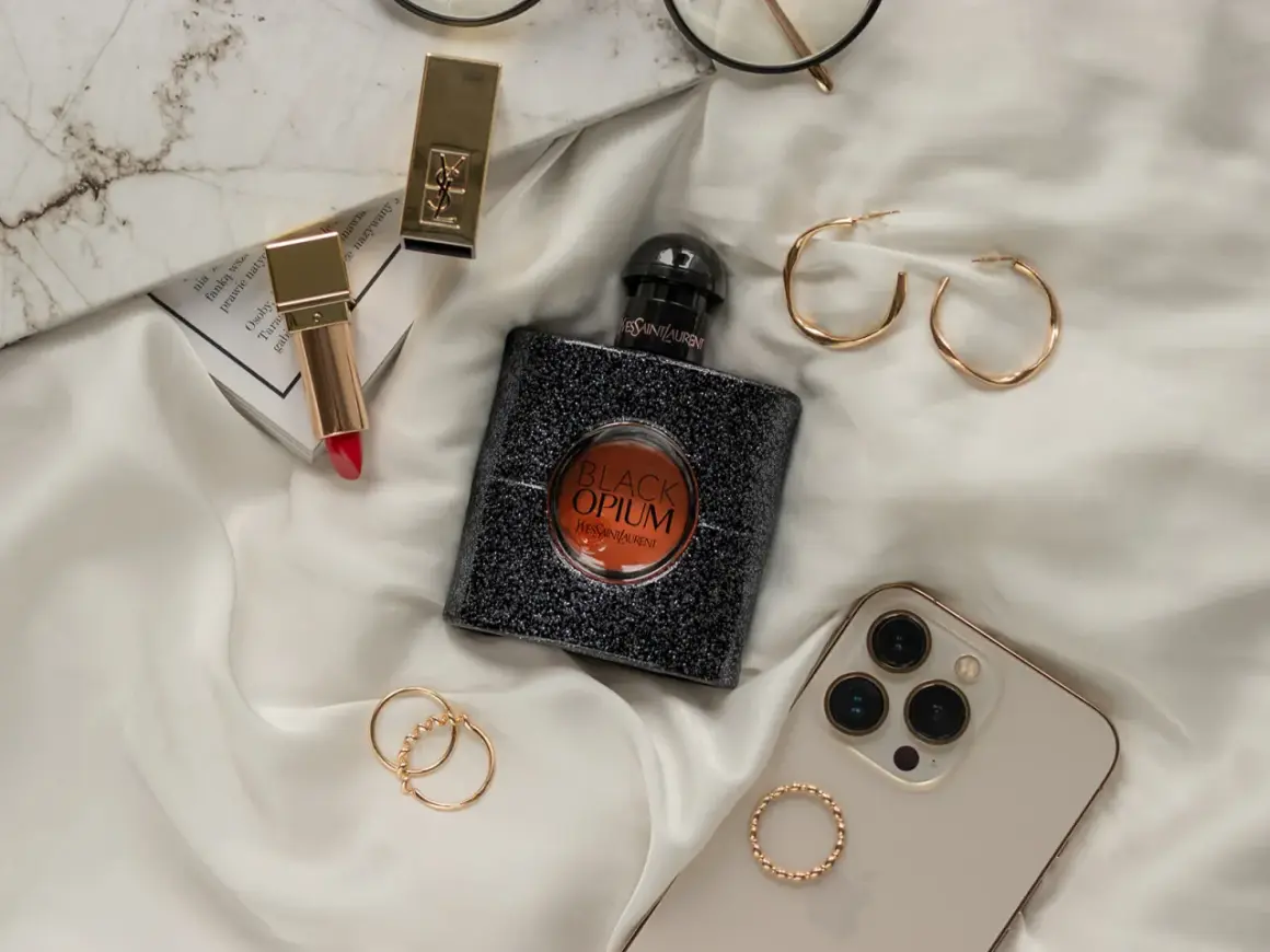 Elegancka kompozycja z flakonem perfum **black opium**, złotą szminką, pierścionkami i telefonem.
