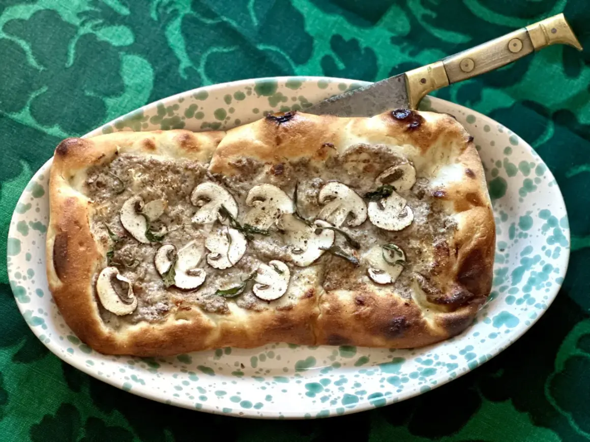 En gyllene pinsa med svamp och örter, redo att skäras med en kniv. Ett perfekt pinsa recept för en mysig kväll.