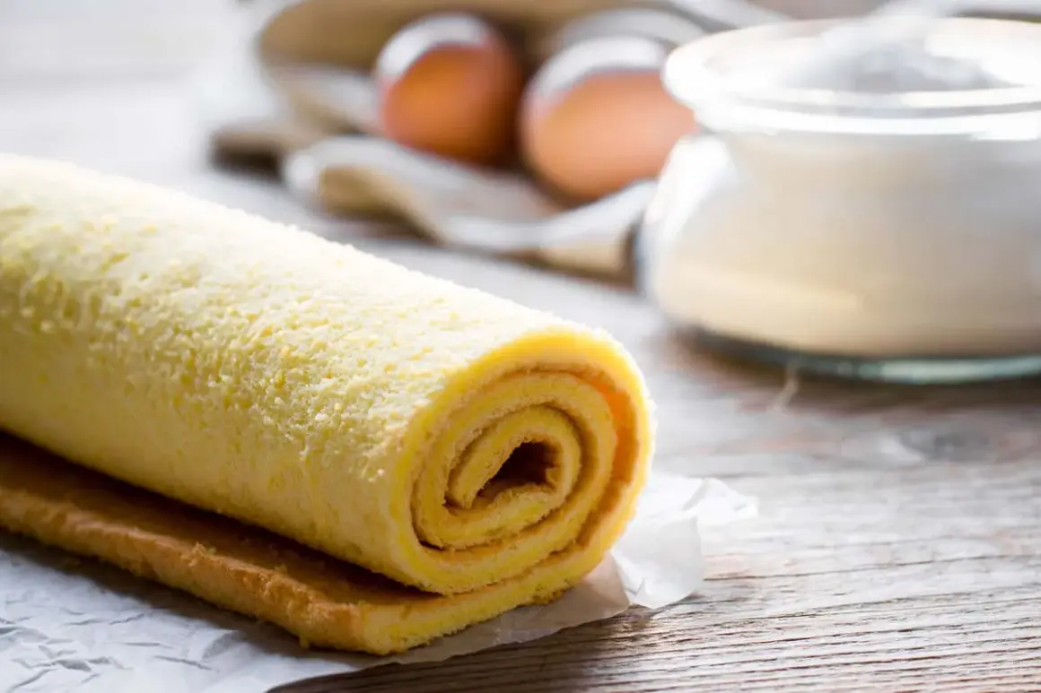 Rotolo di pasta biscotto per torte a strati, pronto per essere farcito. Sullo sfondo, uova e zucchero.
