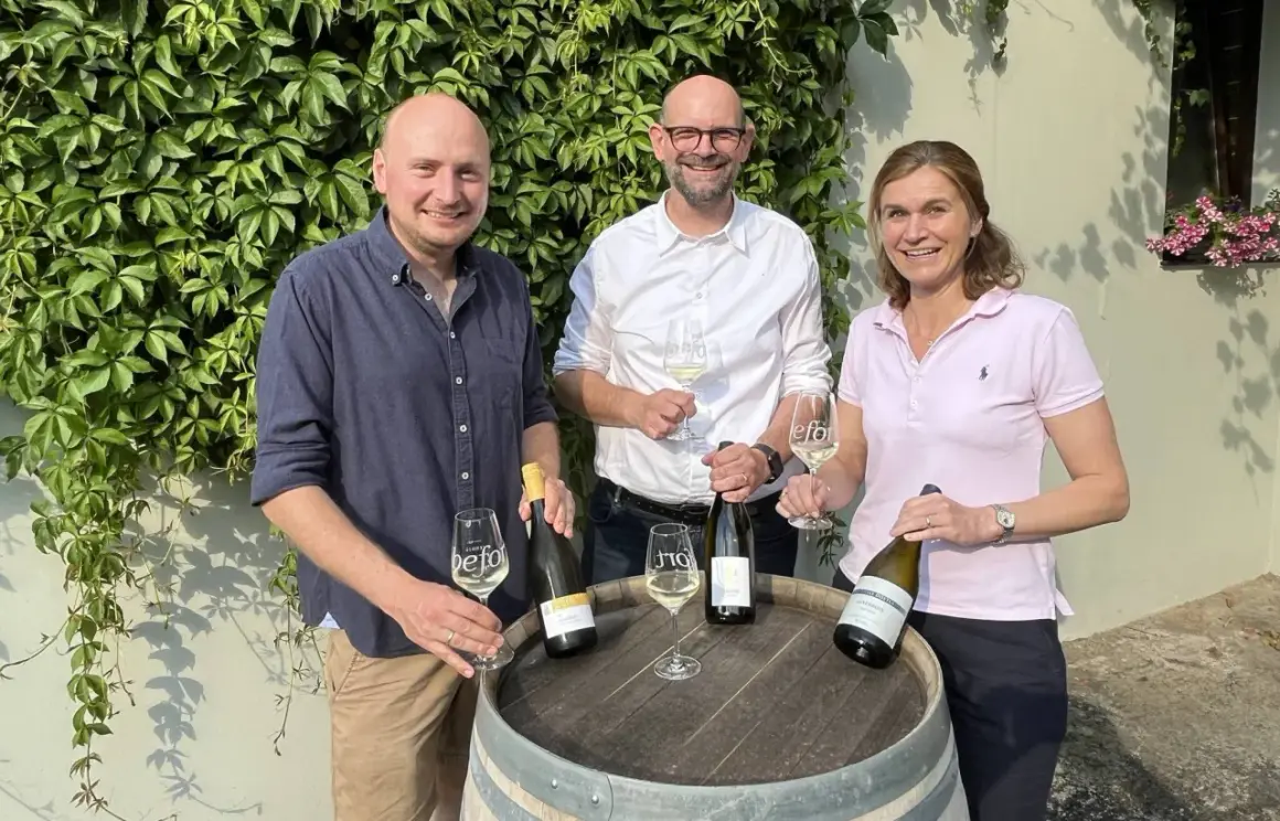Drei Personen mit Wein vor einer Weinrebenwand. Die Flasche ganz rechts trägt das Etikett "Auxerrois".