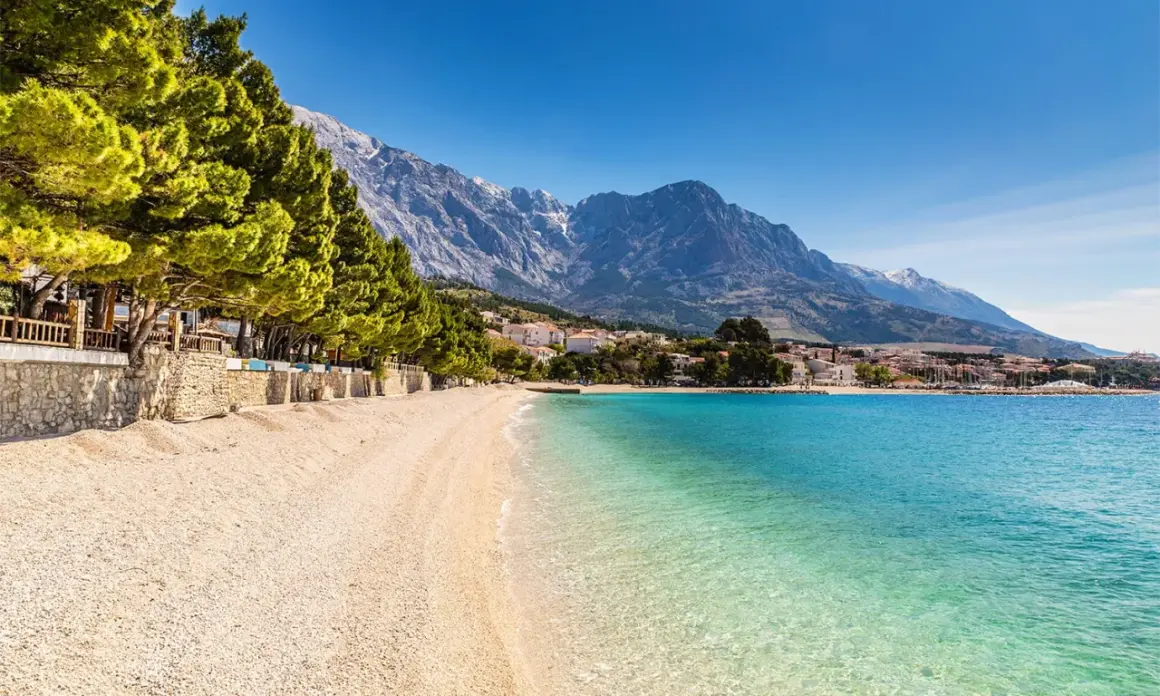 En vacker strand med turkost vatten och en bergskedja i bakgrunden. En promenadstråk kantas av träd och stenmurar, perfekt för en avkopplande stund vid makarska strandpromenad.
