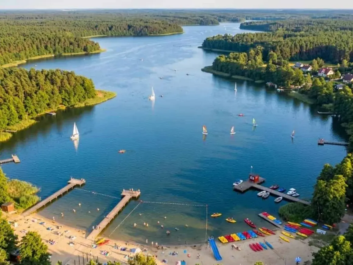 Malownicze jezioro Kałębie z żaglówkami, kajakami i plażą pełną wypoczywających ludzi.