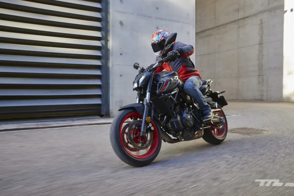 Motociclista con casco y chaqueta roja y gris conduce una Yamaha MT-07 negra con detalles rojos por una calle urbana.