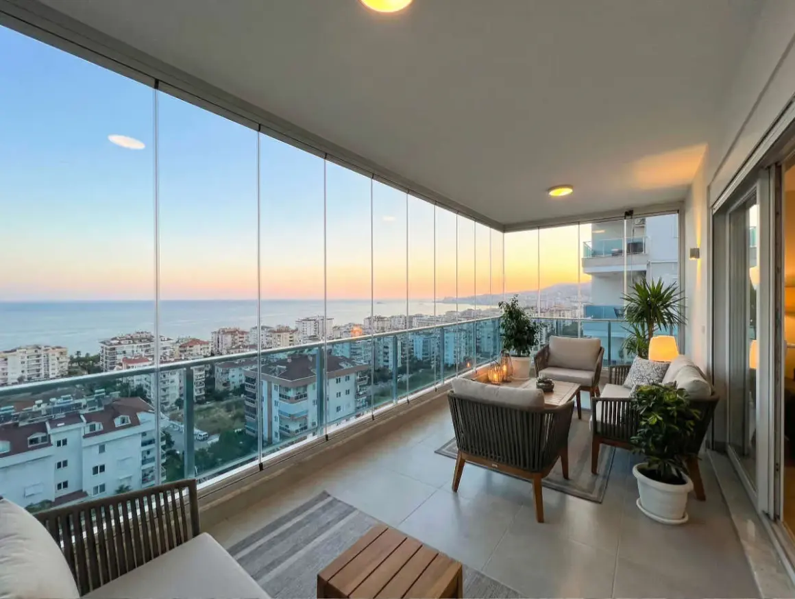 Terraza moderna con cortinas de cristal, muebles de exterior y vistas al mar al atardecer.