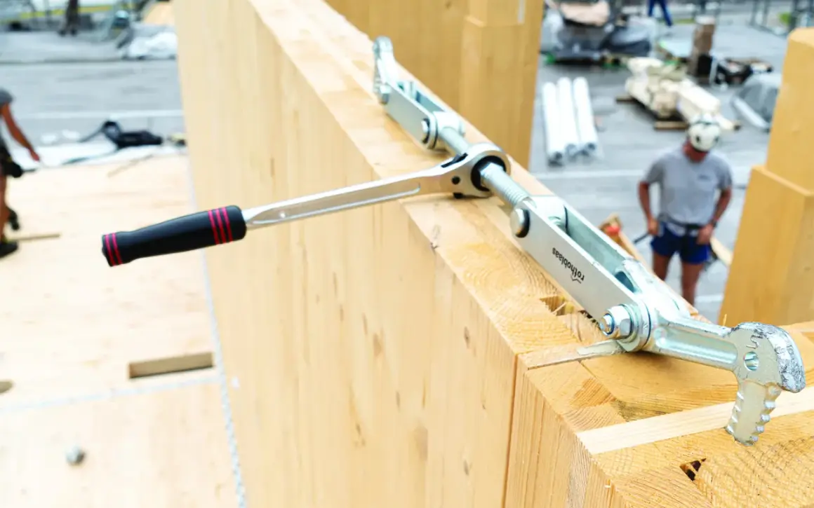 Un outil de serrage métallique, faisant partie d'une liste d'outils de charpentier, est utilisé sur de grosses poutres en bois.