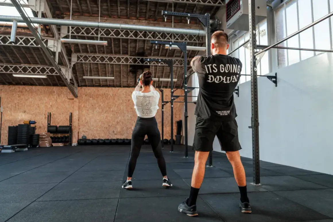 Para osiągnięcia dobrych wyników w crossfit, jak często trenować? Para ćwiczy w siłowni, kobieta z kettlebell, mężczyzna w koszulce "It's Going Down".