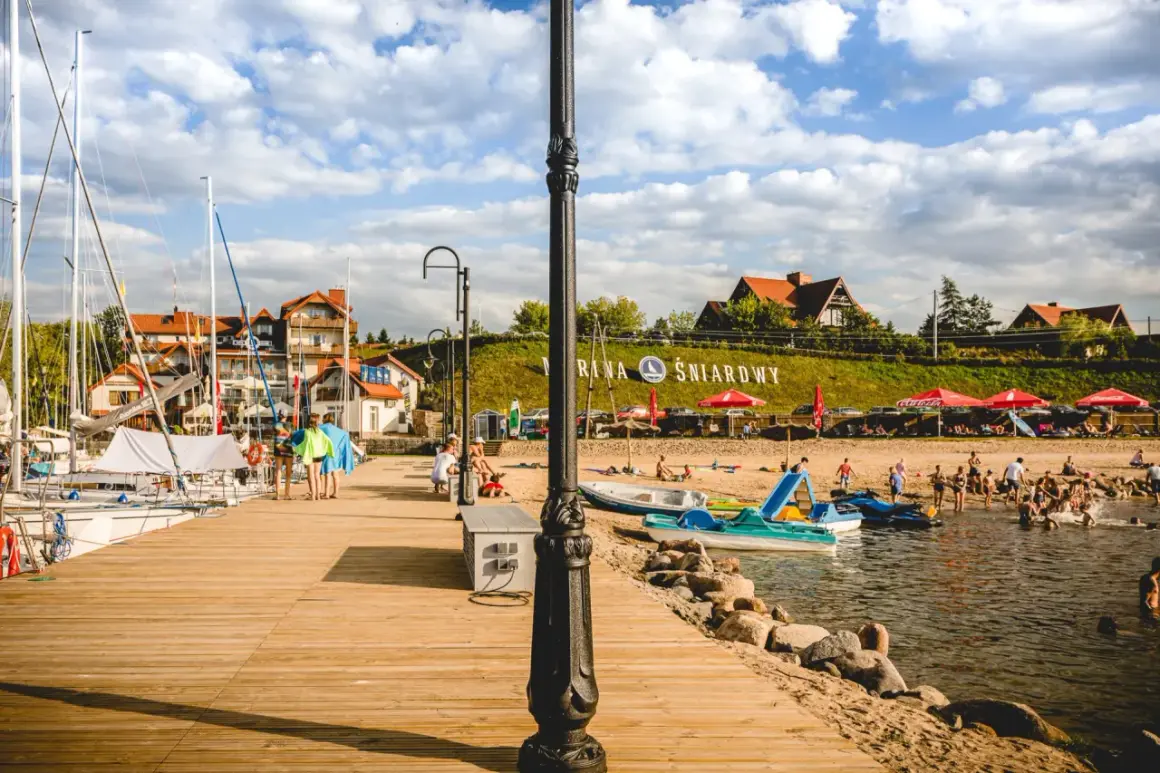 Letnia przygoda w krainie tysiąca jezior. Drewniane molo, żaglówki, plaża z ludźmi i budynki nad jeziorem.