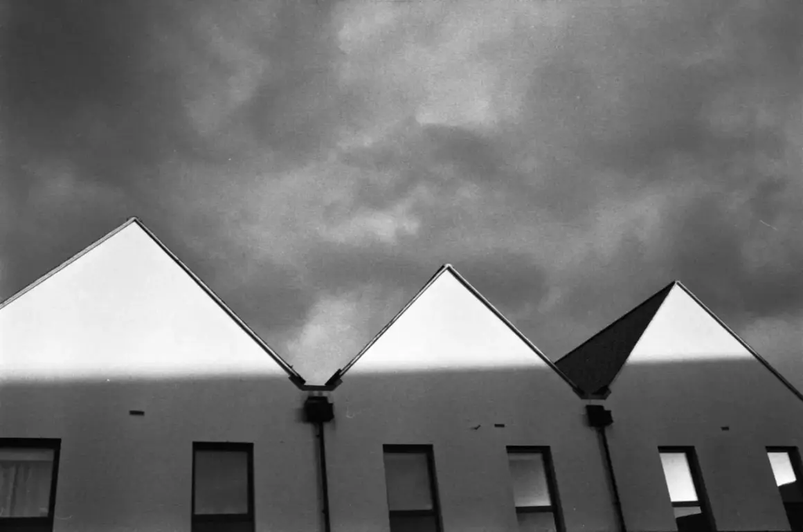 Row of modern houses with sharp, triangular roofs against a dramatic, cloudy sky. The light creates a stark contrast, highlighting the architectural lines, a testament to careful stand development.
