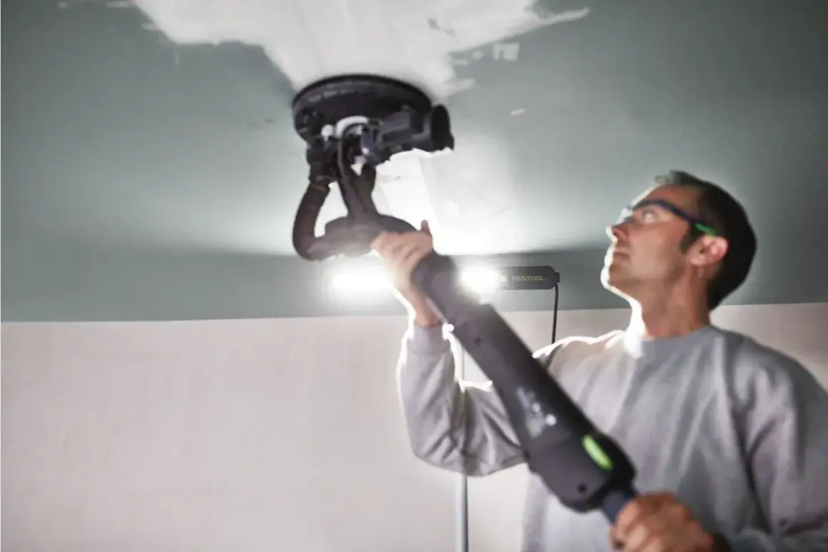 Un homme utilise une ponceuse de plafond Festool, éclairée par une lampe rasante Festool, pour lisser une surface.