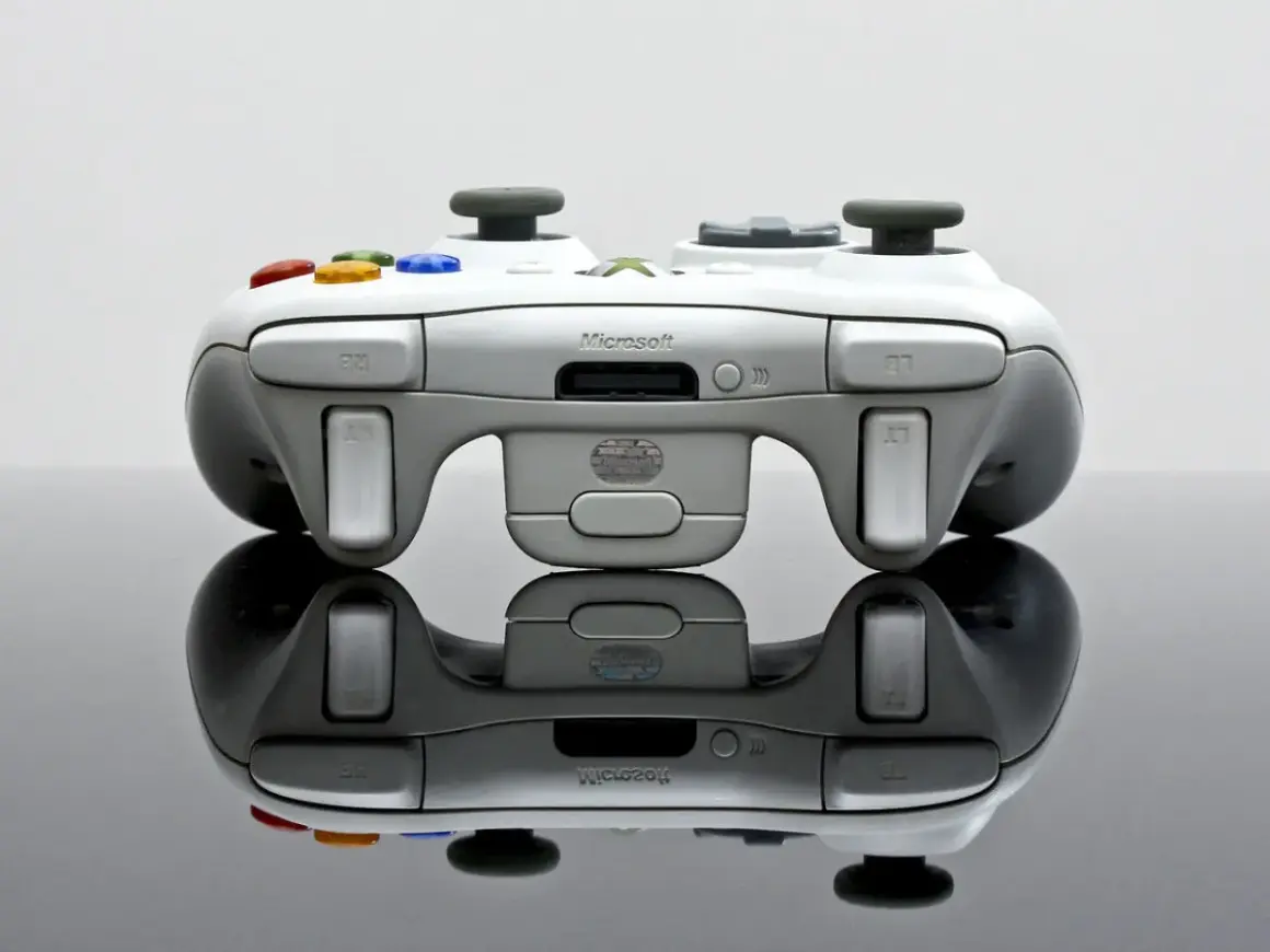 Biały kontroler Xbox 360 z kolorowymi przyciskami, odbijający się od czarnej powierzchni.
