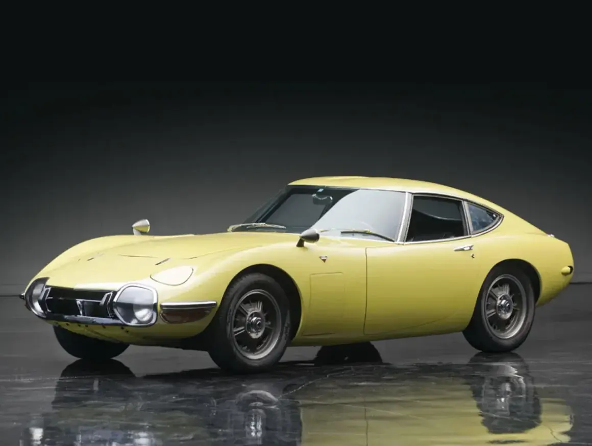 Żółta Toyota 2000 GT z lat 60. prezentuje swoje klasyczne, sportowe linie na błyszczącej podłodze.