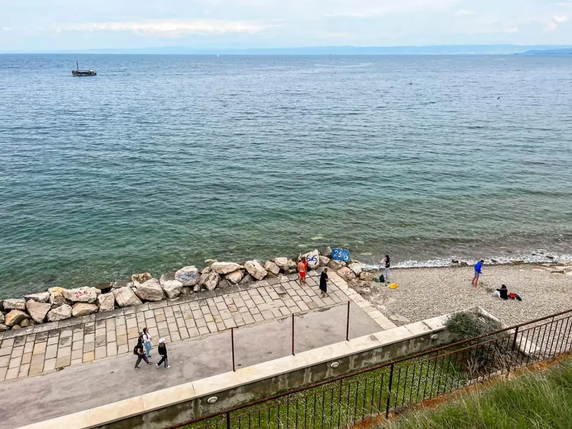 Spacerem po wybrzeżu w Słowenii Piran. Widać łódź na morzu i kilka osób odpoczywających na kamienistej plaży.