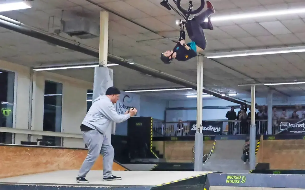 BMX-Fahrer vollführt einen Salto in der Luft in der Wicked Woods Wuppertal. Ein Mann filmt den Stunt.