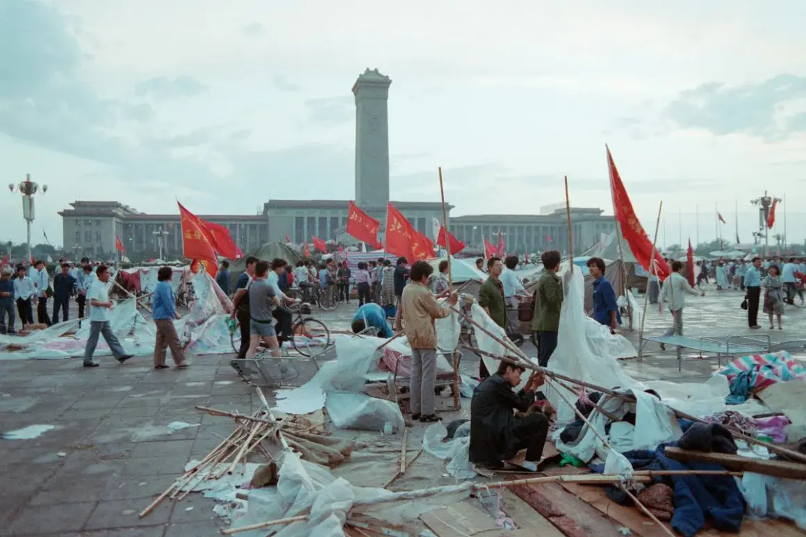 Ludzie na placu Tiananmen, czerwone flagi powiewają, budynki w tle, chaos i prowizoryczne namioty.
