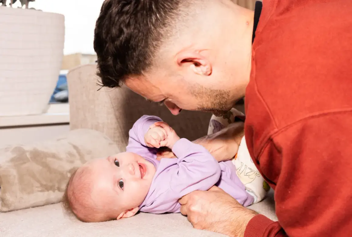 Ein Vater spielt liebevoll mit seinem 4. sprung baby auf einer Couch. Das Baby trägt einen lila Pullover und lächelt.