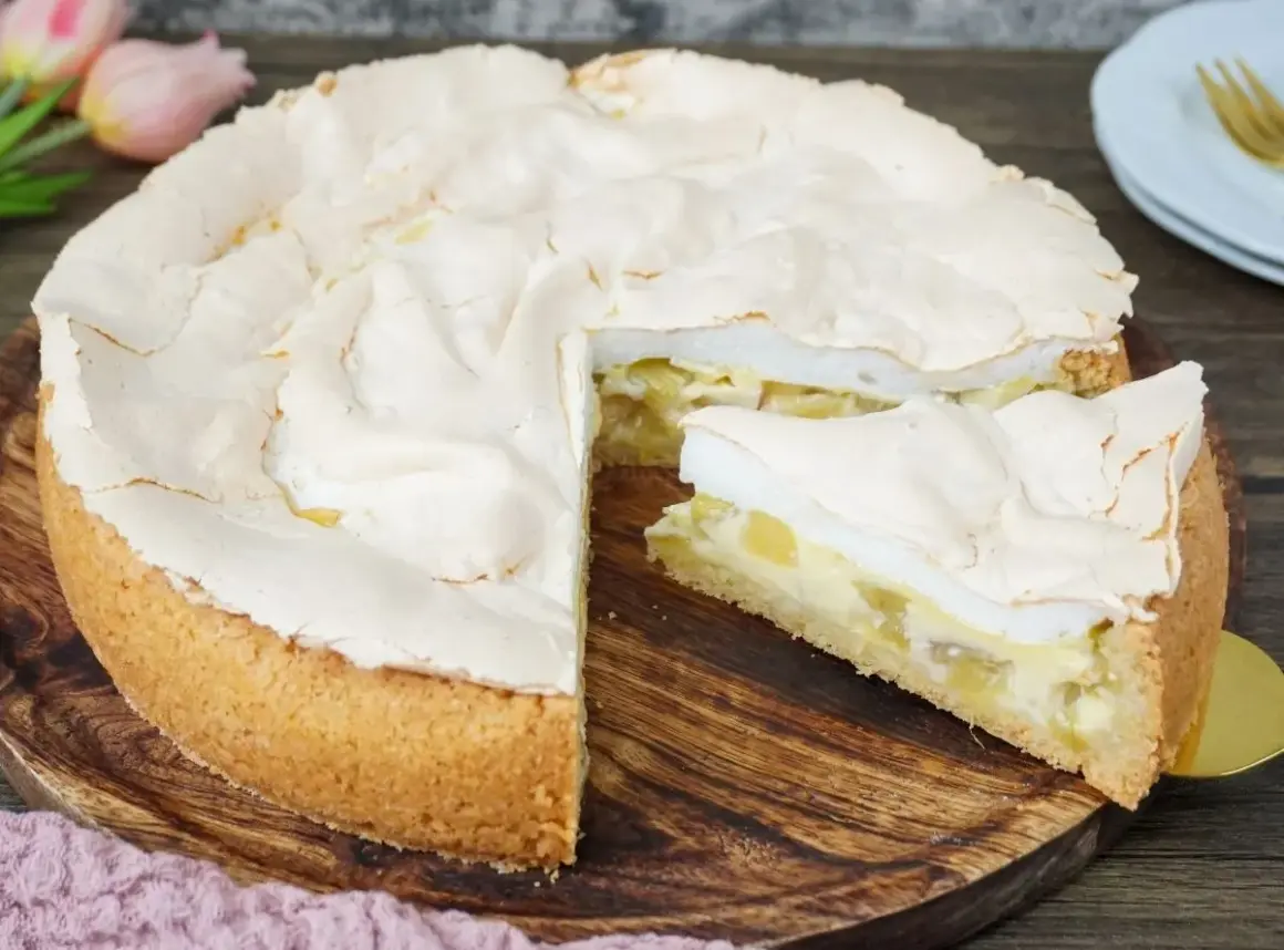 Ein saftiger Rhabarberkuchen mit Baiserhaube, ein Stück davon ist angeschnitten und zeigt die cremige Füllung.