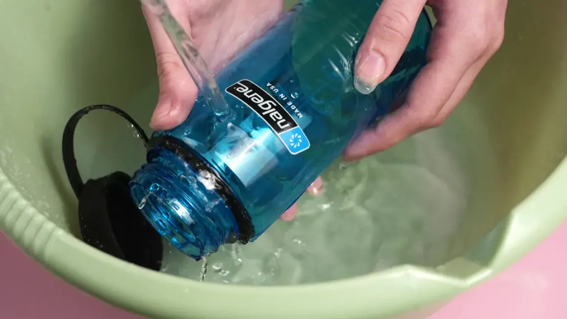 Hände spülen eine blaue Nalgene-Flasche mit kochendem Wasser in einer grünen Schüssel.