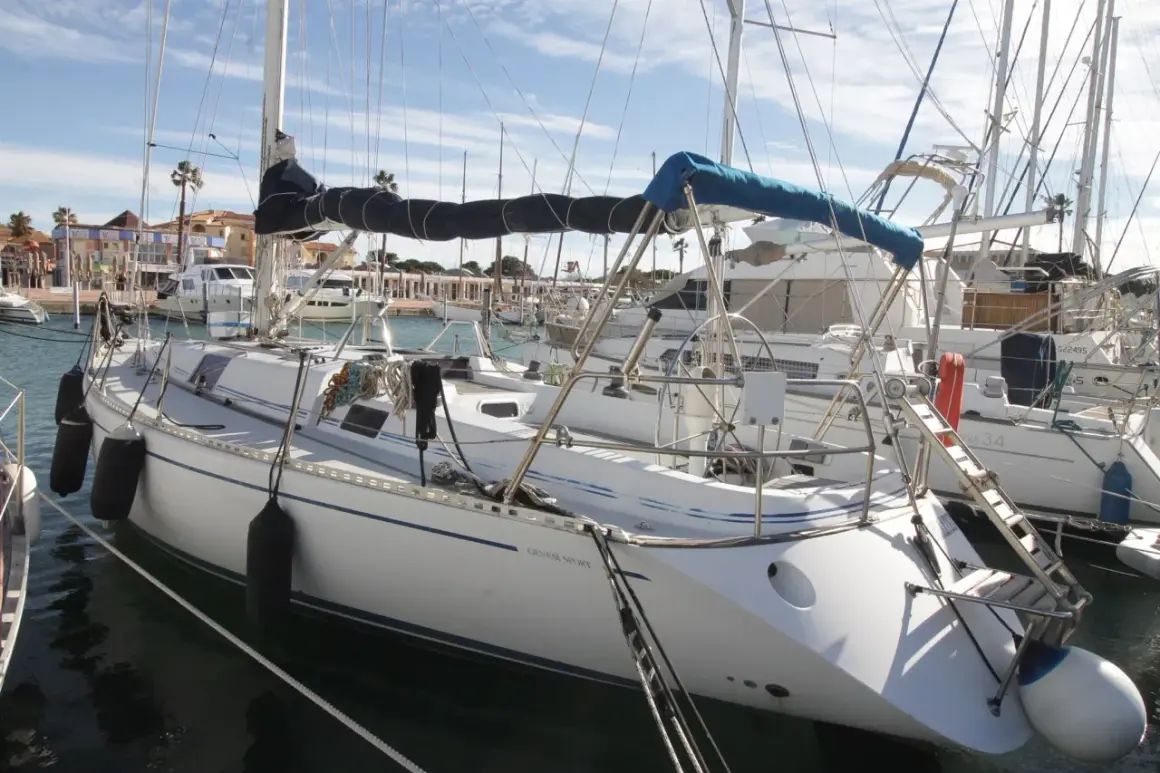 Barche a vela ormeggiate in un porto, con la scritta "Genesi Sport" su una di esse. Cielo azzurro con nuvole.