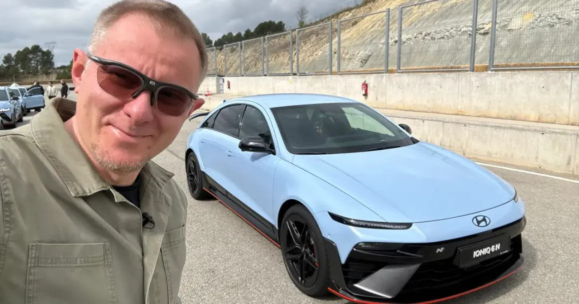 Mężczyzna w okularach przeciwsłonecznych pozuje do selfie z błękitnym Hyundaiem IONIQ 6 N na torze wyścigowym.