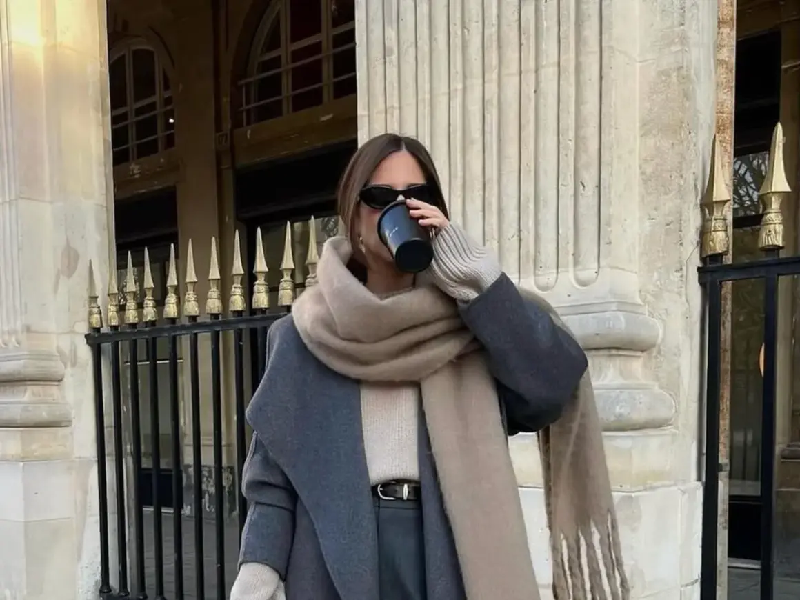Stilvolle Frau trinkt Kaffee, kombiniert grau und beige mit einem Oversize-Schal und Mantel.