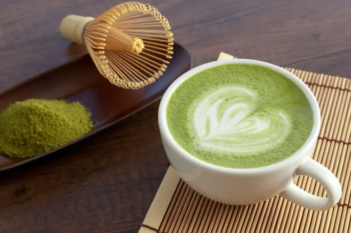 Tazza di matcha latte con schiuma decorata a forma di cuore, accanto a polvere di tè verde e frusta tradizionale.