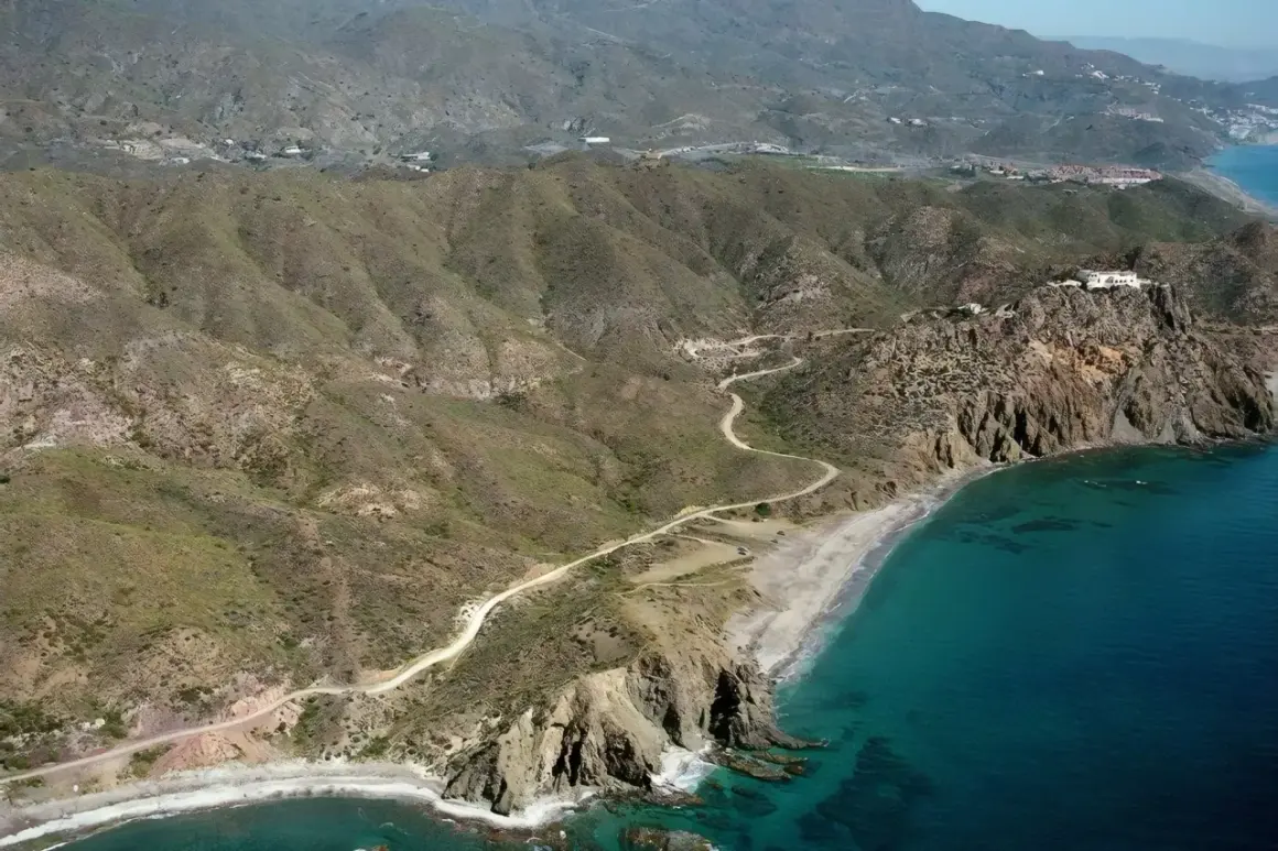 Vista aérea de la playa el lance, con su arena dorada, aguas turquesas y acantilados rocosos. Un camino serpentea por las colinas áridas.