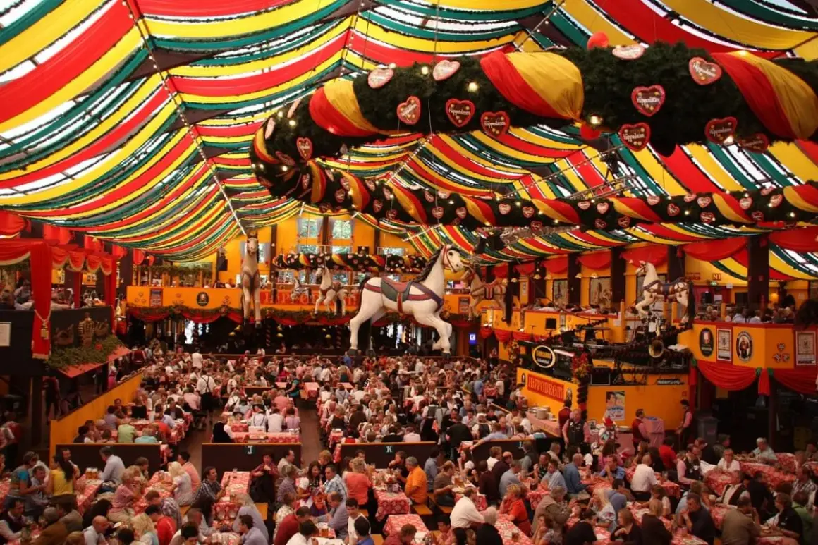 Un tendone colorato ospita la vivace **movida a Monaco di Baviera**. Migliaia di persone siedono ai tavoli, sotto decorazioni a forma di cuore e cavalli da giostra.