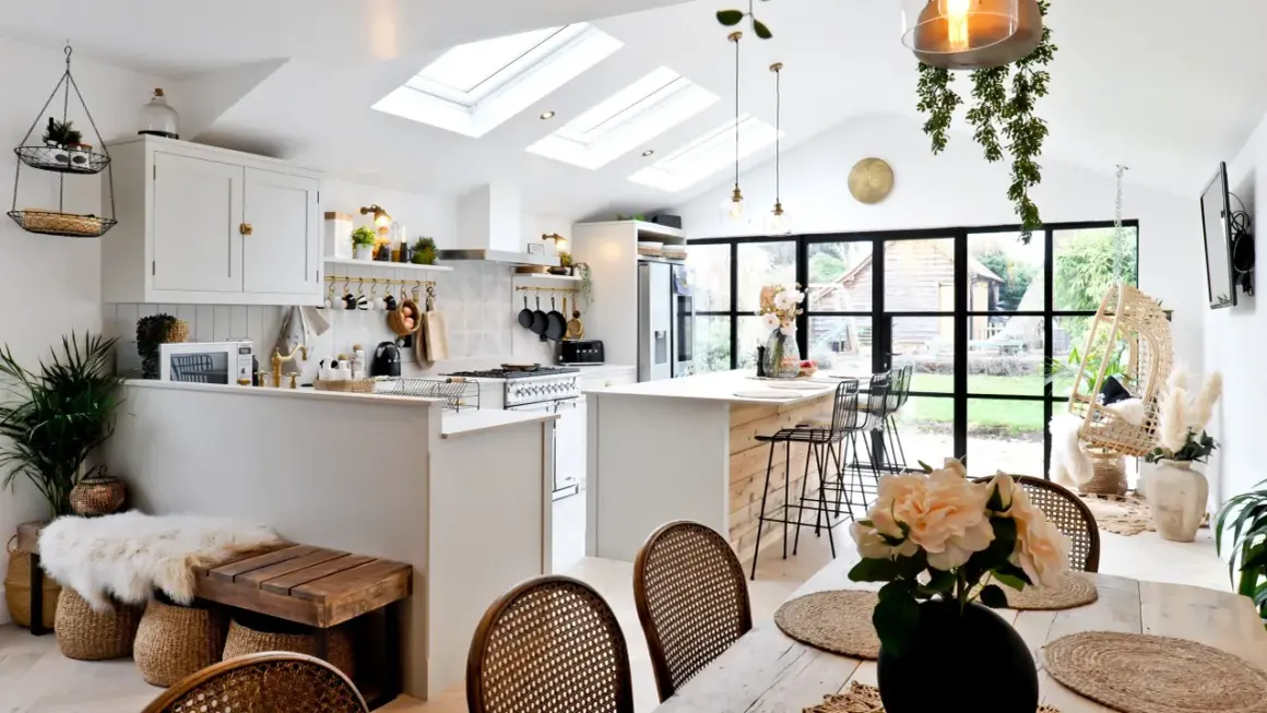 Jasna kuchnia w stylu modern farmhouse z wyspą, białymi meblami i drewnianymi akcentami. Dachowe okna wpuszczają mnóstwo światła.
