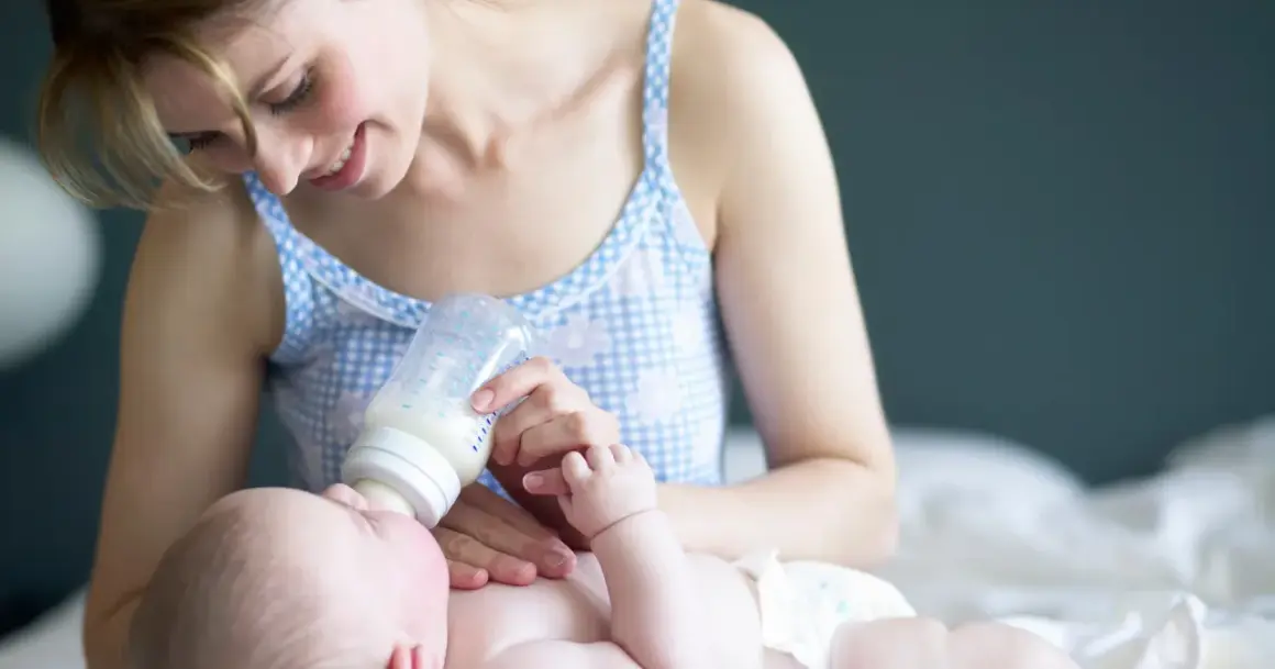 Uśmiechnięta mama karmi butelką noworodka. Warto wiedzieć, ile powinien jeść dwutygodniowy noworodek, by zapewnić mu prawidłowy rozwój.