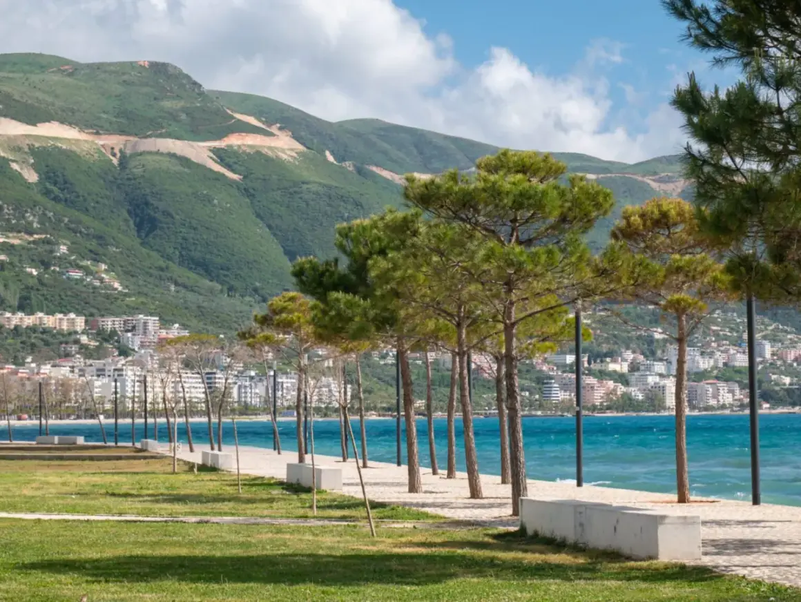 Wlora, Albania: nadmorska promenada z sosnami, turkusowe morze i góry w tle.