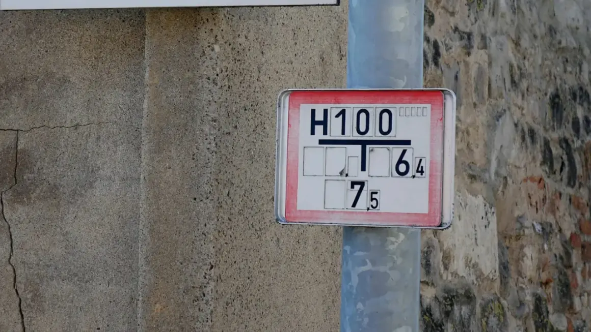 Nahaufnahme eines Hydrantenschilds mit der Aufschrift "H100 T 6,4 7,5". Das Schild ist an einem Metallpfosten befestigt, daneben eine raue Betonwand.