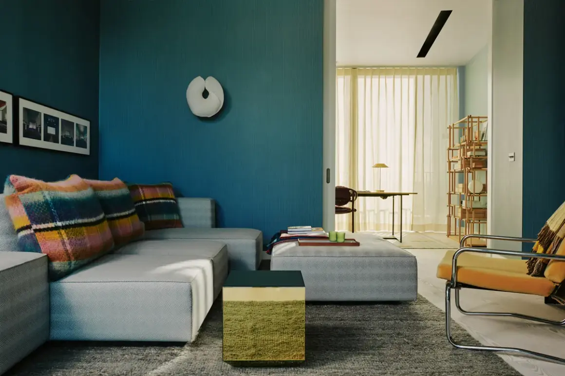 Soggiorno moderno con divano grigio, cuscini colorati e pareti blu intenso. Un tavolino cubico verde e oro aggiunge un tocco di vivacità.