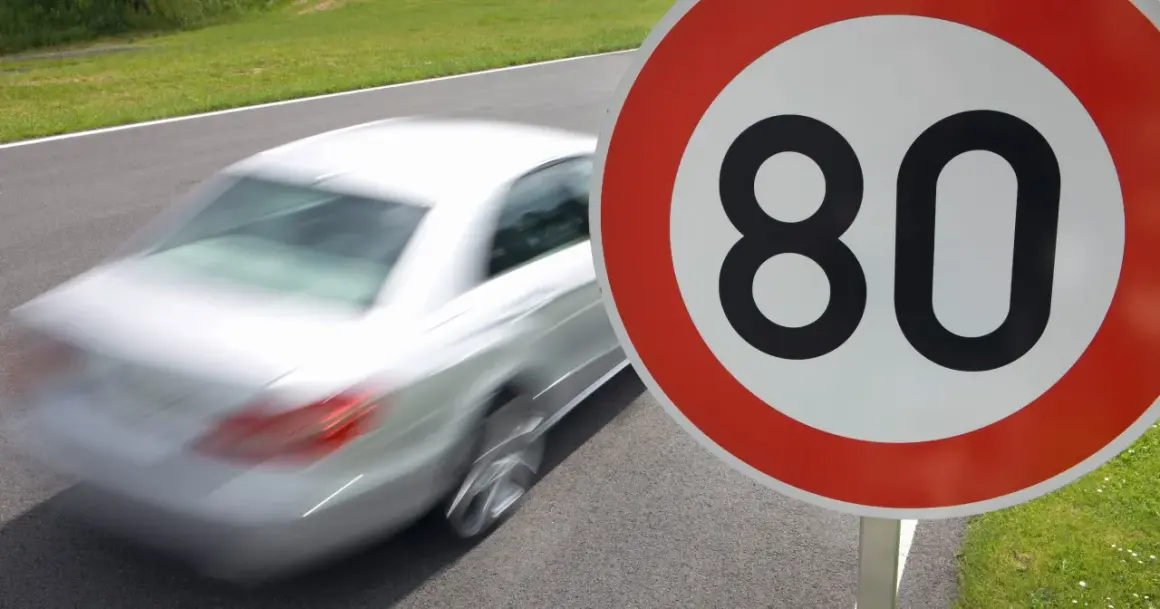 Znak ograniczenia prędkości do 80 km/h. Biały samochód pędzi po drodze, przypominając o przepisach ruchu drogowego i ograniczeniach prędkości w Polsce.