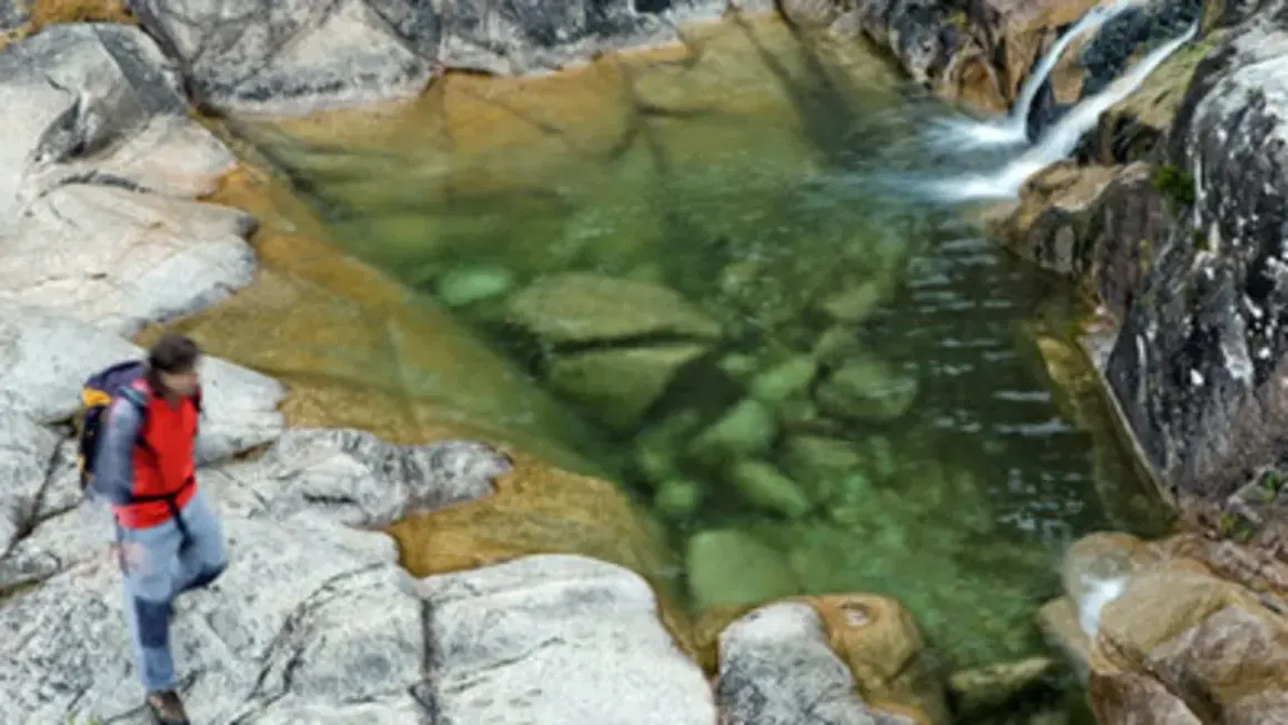 Senderista se detiene junto a un **río caldo** de aguas cristalinas y rocas musgosas.