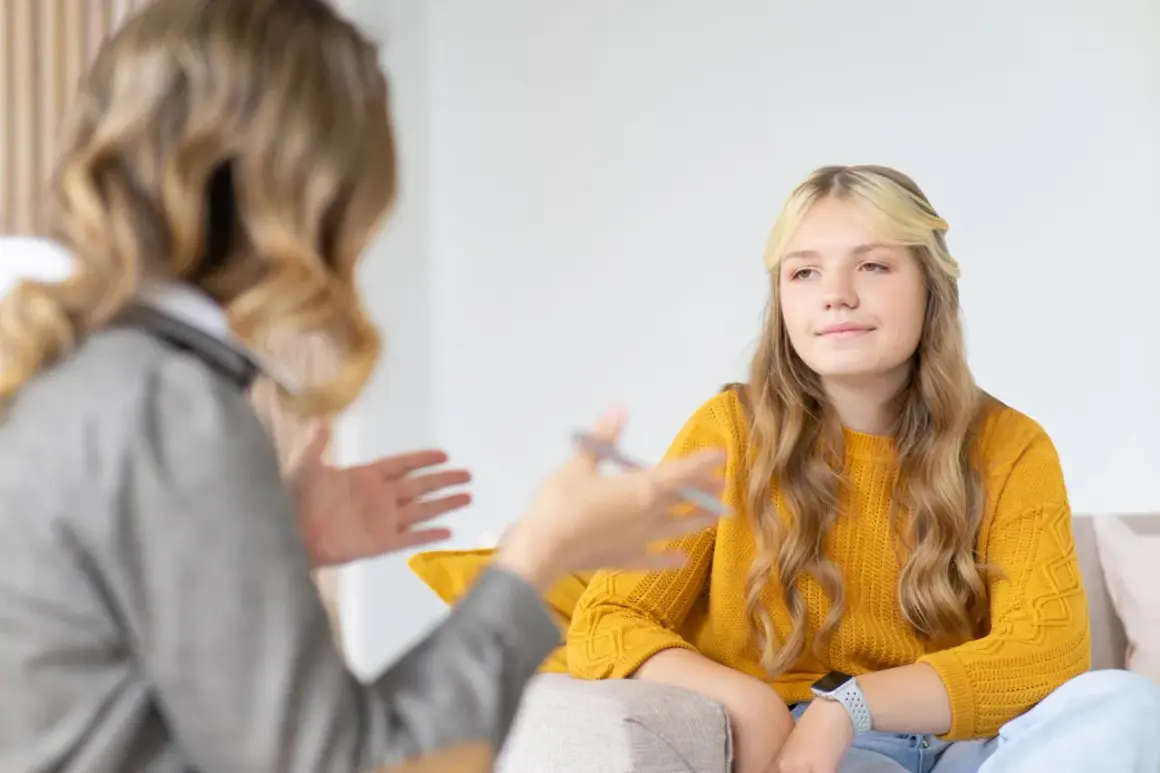Nastolatka w żółtym swetrze podczas terapii. Rozmowa z terapeutką, która gestykuluje.