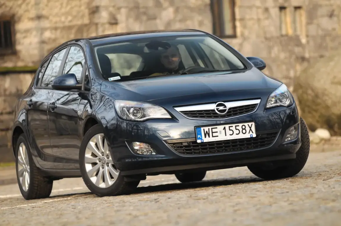 Ciemny Opel Astra J z włączonymi światłami do jazdy dziennej, zaparkowany na kamienistej drodze.