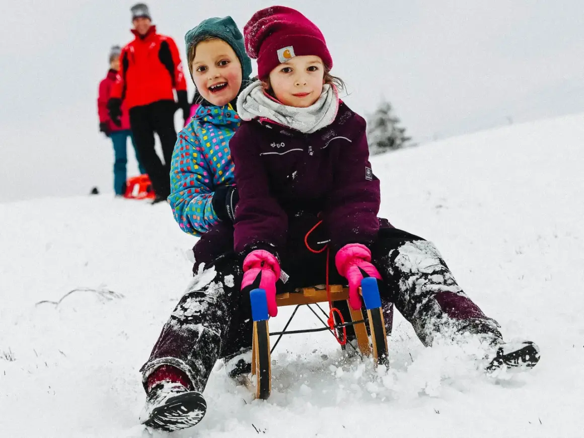 Zwei Kinder rodeln lachend einen verschneiten Hang im Bergischen Land hinunter.
