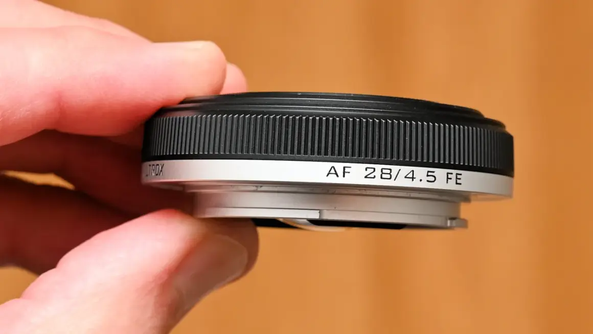 A hand holds a compact, black and silver pancake lens, labeled "AF 28/4.5 FE".