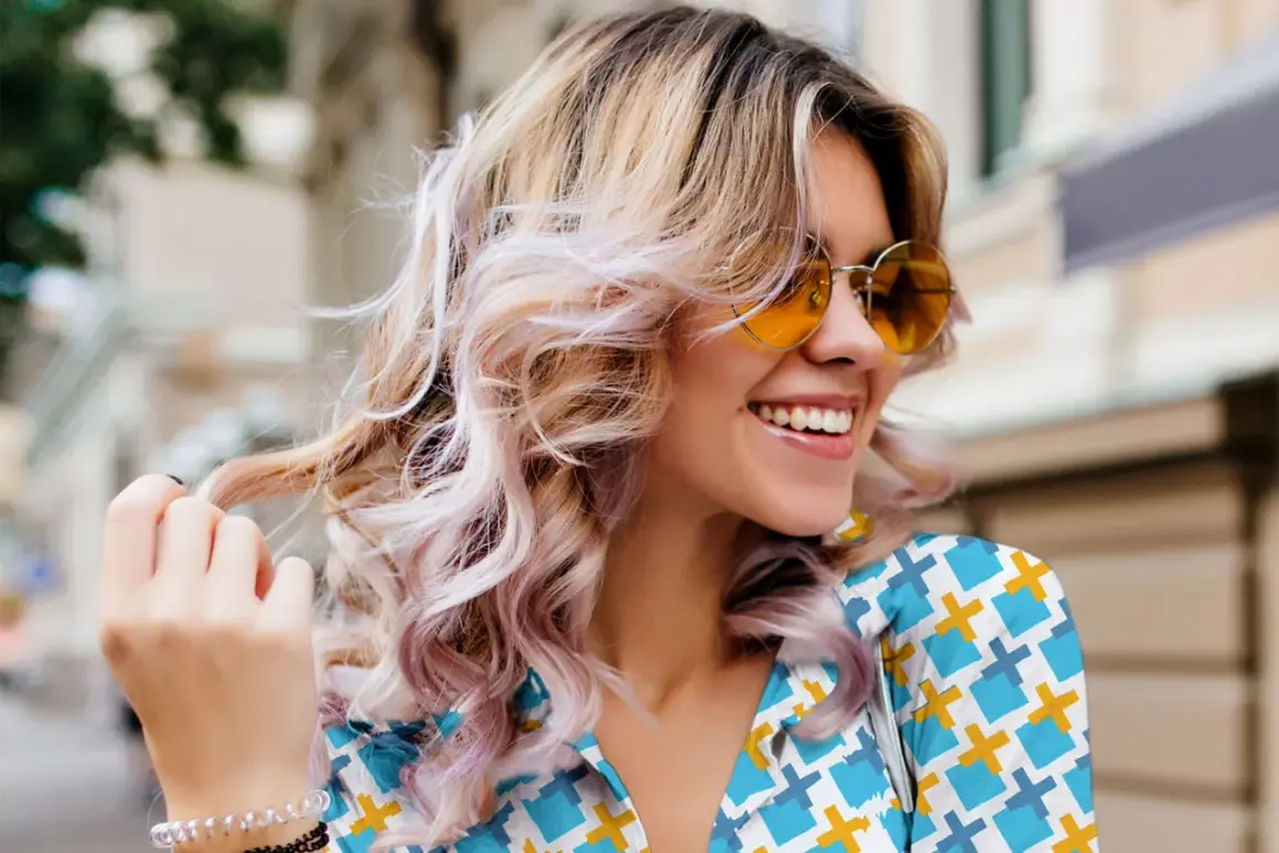 Mujer sonríe con cabello rubio ondulado y mechas rosas, luciendo gafas de sol naranjas. Un ejemplo de cómo reparar el pelo dañado con estilo.