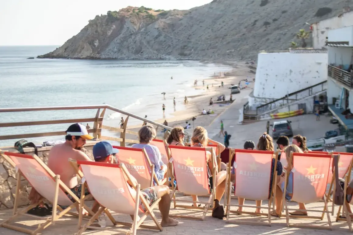 Gente relajándose en tumbonas con vistas a la playa de Burgau. El mar azul y la costa rocosa crean un paisaje idílico.