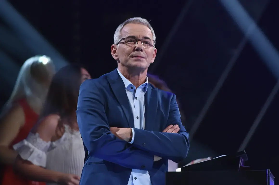 Robert Janowski w garniturze, z rękami skrzyżowanymi na piersi, na tle sceny. Czy to właśnie powód, dlaczego Robert Janowski naprawdę odszedł z Jaka to melodia po latach?