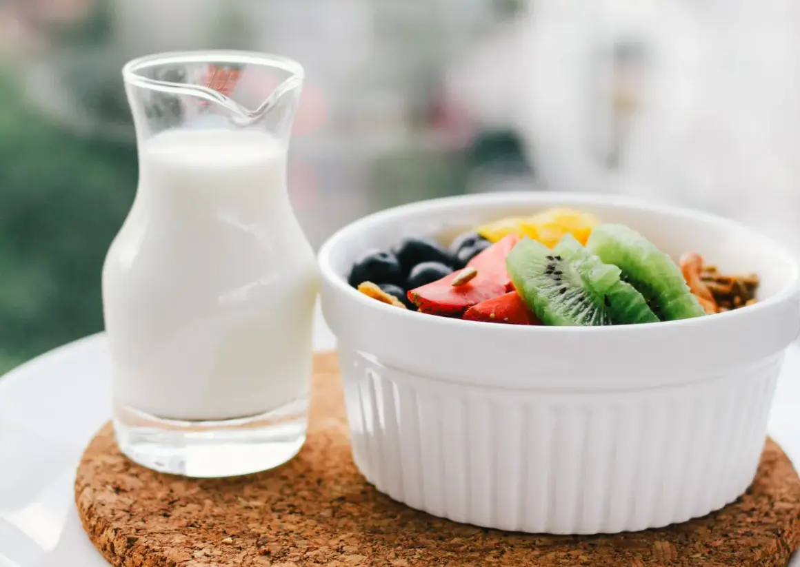 Szklanka mleka obok miseczki z owocami i granolą.